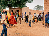 Katsina, Nigeria - 17. Juli 2024: Blick auf Kinder und Erwachsene, die sich im sandigen Hof eines Gebäudes mit Lehmwänden versammeln, wo leuchtende Farben mit den gedämpften Tönen der Erde kollidieren.