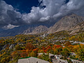 Blick auf herbstliche, in feurigem Rot und Gold leuchtende Bäume inmitten eines Dorfes, eingerahmt von hohen Bergen und einem dramatischen, wolkenverhangenen Himmel, Hunza Nagar, Gilgit Baltistan, Pakistan.