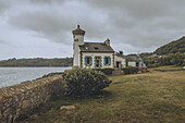 View of the quaint The Phare de Nantouar standing proudly on a grassy hill by the tranquil sea under a cloudy sky, its white walls contrasting with the landscape, Louannec, Bretagne, France.