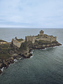 Aerial view of Fort la Latte standing resilient against the crashing waves, its stone walls a stark contrast to the turbulent sea, Plévenon, Bretagne, France.