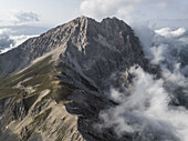 Luftaufnahme des zerklüfteten, sonnenverwöhnten Gran-Sasso-Gipfels, der durch ein Meer aus ätherischen Wolken hindurchragt, ein Tanz von Licht und Schatten auf Stein, Gran Sasso, Abruzzen, Italien.