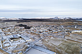 Aerial view of the stark, snow-dusted town nestled near the dark, rugged lava fields, a striking contrast against the serene, distant mountains, Grindavik, Grindavíkurbær, Iceland.