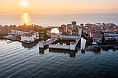Aerial view of the Scaliger Castle, a medieval fortress surrounded by shimmering waters, glows warmly under the setting sun, casting long shadows across the ancient walls, Sirmione, Lombardy, Italy.