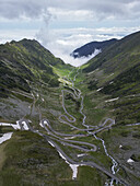 Aerial view of the winding Transfagarasan road meandering through a beautiful mountainous landscape with rugged peaks and green valleys, Cartisoara, Sibiu, Romania.