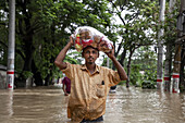 Satkania, Bangladesh - 09 August 2023: View of a man wading through floodwaters, carrying a large bag of colorful items overhead, amidst submerged streets and trees.