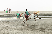 Cox's Bazar, Bangladesch - 29. November 2022: Blick auf ein Kind, das am Strand von Cox's Bazar auf einem weißen Pferd reitet, dessen Schwanz hoch fliegt, inmitten der gedämpften Töne von Sand und Himmel.