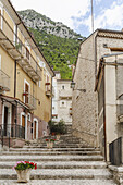 Blick auf sonnenüberflutete Steintreppen, die durch eine enge Gasse führen, flankiert von Gebäuden mit bunten Fassaden unter einem weiten Himmel, Pacentro, Abruzzen, Italien.