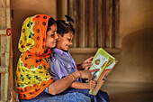 Bogura, Bangladesch - 15. Dezember 2019: Blick auf eine Mutter und ein Kind, die in sanftes Licht getaucht sind und vor dem Hintergrund eines einfachen Hauses ein buntes Buch lesen.