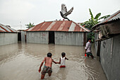 Kurigram, Bangladesch - 21. Juni 2022: Blick auf Kinder, die durch die Fluten waten, eine deutliche Erinnerung an die Macht der Natur über die Wellblechhäuser, während eine Taube die Flucht ergreift.