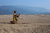 Chattogram, Bangladesh - 27 January 2019: View of a worker sifting sand on a vast, sun-drenched beach, with hazy mountains rising in the distance, creating a scene of labor against a serene backdrop.