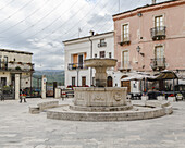 Blick auf einen steinernen Brunnen, der stolz auf einer sonnenüberfluteten Piazza steht, umgeben von Gebäuden mit pastellfarbenen Fassaden, unter einem Himmel mit vereinzelten Wolken, Pacentro, Abruzzen, Italien.