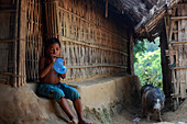 Mirsharai, Bangladesh - 21 October 2019: View of a young boy sitting outside a bamboo hut, blowing a blue balloon, as a pig forages nearby, capturing a moment of simple rural life.