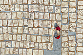 Bogura, Bangladesch - 30. Juli 2025: Luftaufnahme von fein säuberlich angeordneten, trocknenden Lebensmittelquadraten, die neben einer Figur in leuchtendem Rot lange Schatten werfen und ein Mosaik aus Texturen und Farbtönen bilden.