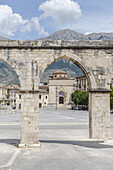 Blick auf einen alten steinernen Torbogen, der eine sonnenüberflutete Piazza mit einem kuppelförmigen Gebäude umrahmt, hinter dem sich schroffe Berge gegen einen hellen Himmel abzeichnen, Sulmona, Abruzzen, Italien.