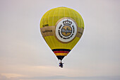 Chambley, France - 30 July 2025: View of a bright yellow hot air balloon emblazoned with a beer logo floats against a pale, overcast sky.