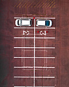 Aerial view of two white cars parked neatly in a red parking lot with white lines and directional arrows, Chikusei, Ibaraki, Japan.