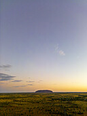 Luftaufnahme des ikonischen Uluru-Monolithen, der sich aus der flachen, trockenen Landschaft unter einem weiten, in Orange- und Blautönen gefärbten Himmel erhebt, Petermann, Northern Territory, Australien.