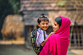 Bogura, Bangladesch - 15. Dezember 2019: Blick auf eine Mutter, die ihr lächelndes Kind mit Schuluniform und Rucksack im Arm hält, vor der Kulisse eines traditionellen ländlichen Hauses.