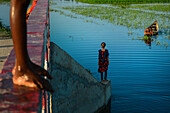 Bogura, Bangladesh - 29 September 2015: View of a young girl in a patterned dress standing at the edge of a concrete structure overlooking the serene, reflective water where a small boat drifts.