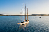 Aerial view of a majestic sailing yacht gliding across the tranquil, deep blue waters, kissed by the golden light of the setting sun, Ermioni, Argolis, Greece.
