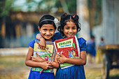 Bogura, Bangladesh - 13 December 2019: View of two girls in blue dresses, beaming with youthful exuberance, embrace each other while holding their schoolbooks tightly.