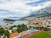Luftaufnahme der Küstenstadt Makarska, Splitsko-dalmatinska županija, Kroatien, eingebettet zwischen der Adria und hoch aufragenden Bergen, ein lebendiger Wandteppich aus roten Ziegeldächern.