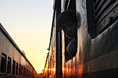 Chittagong, Bangladesh - 02 November 2018: View of a young boy peering from a train window as the golden light of dawn casts long shadows across the carriages.