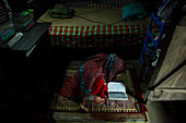 Mirsharai, Bangladesh - 07 May 2020: View of a woman in a vibrant red patterned shawl absorbed in reading on a woven mat, the scene lit by soft light against the room's shadows.