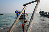Cox's Bazar, Bangladesh - 07 March 2022: View of a boy standing by weathered wooden structures, with fishing boats resting on the glistening shore under a pale sky.