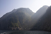 View of dramatic cliffs blanketed in deep green forests rise from the dark waters of Resurrection Bay, shrouded in a mystical haze of sunlight, Kenai Fjords National Park, Seward, Alaska, United States.