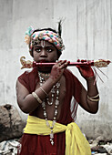Narayanganj, Bangladesh - 23 August 2019: View of a young boy dressed as Krishna, his face painted, playing a flute against a backdrop of textured, muted concrete.