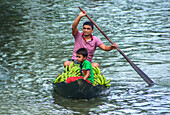 Bogura, Bangladesch - 28. Juli 2017: Blick auf einen Mann und ein Kind, die in einem kleinen, mit leuchtend grünen Bananen beladenen Boot über den Fluss fahren, wobei sich der bedeckte Himmel im Wasser spiegelt.