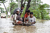 Satkania, Bangladesh - 09 August 2023: View of floodwaters engulfing the streets, reflecting the overcast sky as men navigate the deluge, pushing a bicycle through the murky depths.
