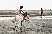 Cox's Bazar, Bangladesh - 29 November 2022: View of a young boy atop a white horse, its mane flowing in the breeze, standing on the sandy beach as the tide gently rolls in.
