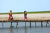 Bogura, Bangladesch - 16. Februar 2018: Blick auf zwei Frauen, von denen eine ein Kind trägt, die eine rustikale Bambusbrücke über ein ruhiges Gewässer unter einem klaren Himmel überqueren.