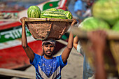 Dhaka, Bangladesh - 08 April 2023: View of a man balancing a basket of vibrant watermelons on his head near the bustling riverfront, a scene of daily commerce and vibrant hues.