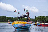 Chattogram, Bangladesh - 19 September 2023: View of vibrant fishing boats adorned with national flags bobbing gently on the water, reflecting the overcast sky and lush green shoreline.