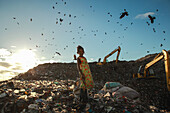 Chattogram, Bangladesh - 19 September 2022: View of a young girl amidst a sprawling landfill, with a flock of birds soaring above and heavy machinery looming in the background.