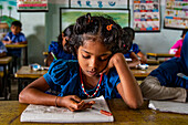 Bogura, Bangladesh - 14 December 2019: View of a young girl with dark curly hair deeply engrossed in her studies at a wooden desk in a sunlit classroom.