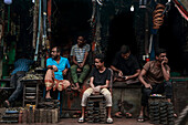 Dhaka, Bangladesh - 09 November 2019: View of men seated in the dimly lit Dhaka metal market, surrounded by a labyrinth of hanging wires and metallic parts.