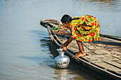 Bogura, Bangladesh - 20 January 2019: View of a young girl in a vibrant floral dress drawing water from a river into a metal jug from a wooden boat.