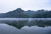 View of still lake waters mirror lush, green hills shrouded in mist, creating a tranquil and ethereal scene, Rangamati, Chittagong Division, Bangladesh.