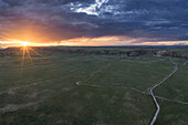 Aerial view of the sun setting over the vast green plain, with a road cutting through the landscape under the dramatic sky, Qinghai Lake, Qinghai, China.