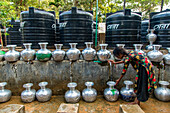 Bogura, Bangladesh - 11 December 2019: View of a girl filling a silver pot with water from a tap against a backdrop of dark water tanks.