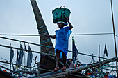 Chittagong, Bangladesh - 28 November 2019: View of a man balancing a green basket, a striking figure against the backdrop of fishing boats.