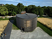 Aerial view of the Chapel of Reconciliation's oval structure, dark wooden facade contrasting with golden fields and Berlin TV Tower in the distance, Berlin, Germany.