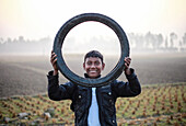 Rangpur, Bangladesh - 01 December 2019: View of a smiling boy holding a tire in a field, framing his face against the soft, hazy horizon.