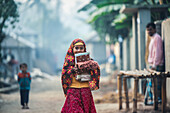 Bogura, Bangladesh - 04 January 2021: View of a young girl with a vibrant, patterned shawl and books smiles amidst the dusty path of rural Bangladesh.