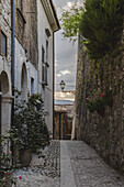 View of an old narrow street, cobblestone path leading to an arched doorway under a vintage street lamp, flanked by weathered stone walls and blooming plants, Pacentro, Abruzzo, Italy.