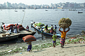 Dhaka, Bangladesh - 29 December 2020: View of vibrant riverbank activity where laborers carry heavy loads amidst boats laden with produce, framed by the city skyline.